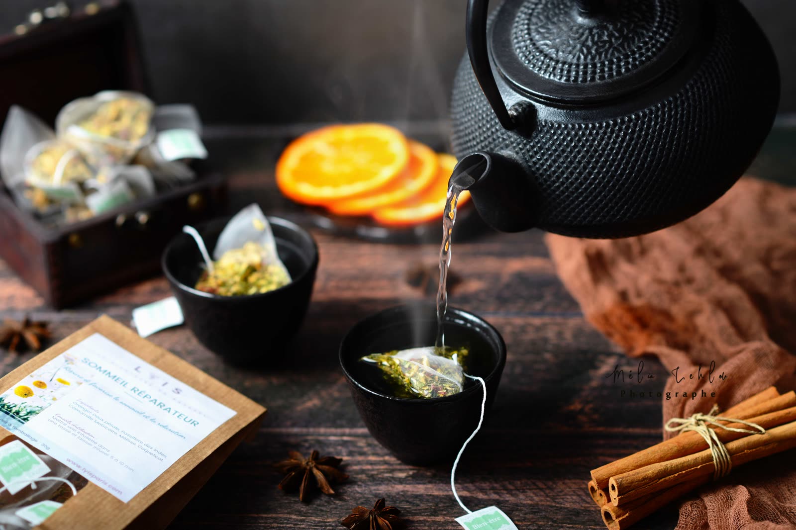 Théière en fonte versant une infusion dans un bol noir, oranges séchées et cannelle